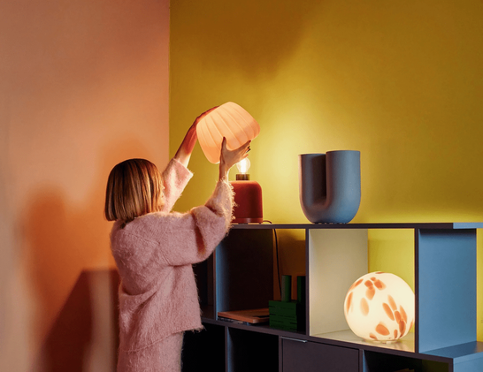 Une personne ajustant un abat-jour coloré sur une lampe, placée sur une étagère moderne avec divers objets de décoration, le tout dans un environnement aux murs aux couleurs vives.