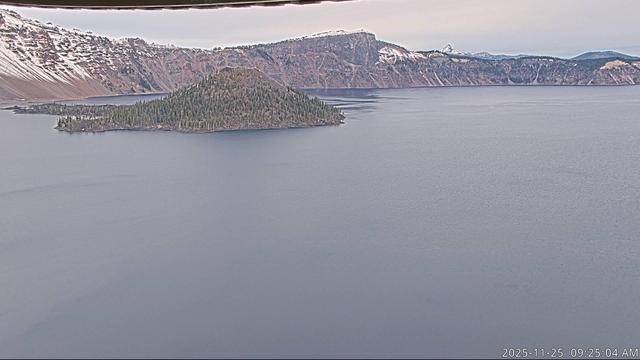 The view of Crater Lake from the Sinnott Memorial Overlook today. Most of the foreground is the lake surface, which is nearly uniformly grey today. Wizard Island provides a touch of color, with green forested slopes. Behind, snow and rock encircle the lake, under a mostly cloudy sky. The camera is back in operation now thanks to an update and upgrade from NPS staff.