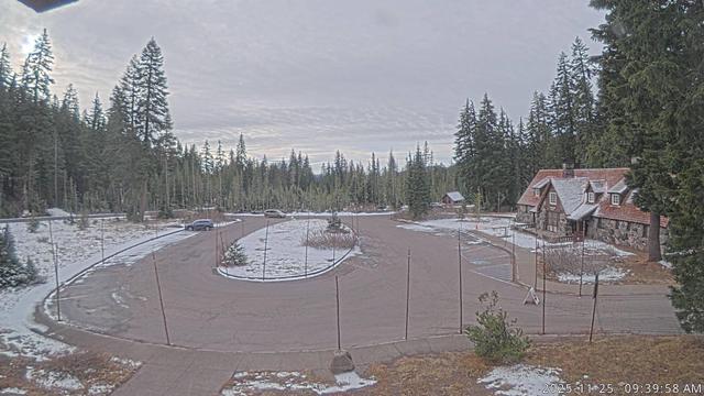 A view of Park Headquarters and the Steel Information Center in Crater Lake National Park today. Much of the image shows the parking area, which is presently generally free of snow. Trees line the horizon, with the information center on the right. Snow poles can be seen at the boundaries of the parking area along with a hint of the sun (on the left) under a mostly cloudy sky. This camera is back in operation now after an update and upgrade by NPS staff.