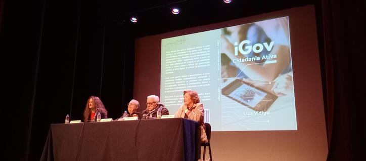 Mesa da apresentação do livro: da esquerda para a direita, Elisabete Carvalho, Luís Vidigal (autor), José Tribolet e Helena Monteiro. Atrás da mesa, a capa do livro "iGov: cidadania ativa" ocupa toda a parede.