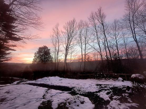 Une photo prise très tôt le matin. 
En avant plan, un terrain plat avec un peu de neige. Au loin, des arbres et derrière les arbres une colline derrière laquelle le soleil se lève. Le ciel est rose et bleu.