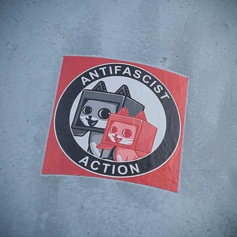 Autocollant antifasciste collé sur un poteau métallique gris
Dans un carré rouge, y'a un gros cercle noir où on peut lire Antifascist Action 
À l'intérieur y'a deux potichats rigolards avec un carton ouvert sur la tête, l'un rouge et l'autre noir