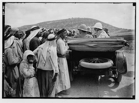 The image depicts a historical moment, likely from the early 20th century given its black and white composition. A group of people is gathered around an open vehicle on what appears to be a rural road or dirt path. The individuals are dressed in traditional Middle Eastern attire such as long robes, headscarves (keffiyeh), turbans (ghutras) and hats which suggest they may belong to the local population.
In contrast, three people wearing safari-style jackets with wide-brimmed hats sit inside the vehicle. These could be British officials or explorers from that era given their distinct style of clothing compared to those outside the car. One individual appears to be in a discussion with someone who is seated and facing them within the vehicle.
The background features hilly terrain, which indicates an arid landscape typical for regions like Palestine during this period. The image captures a moment possibly related to British colonial administration or exploration as Sir Herbert Samuel was indeed visiting Palestinian villages at that time according to caption information provided by archive source.