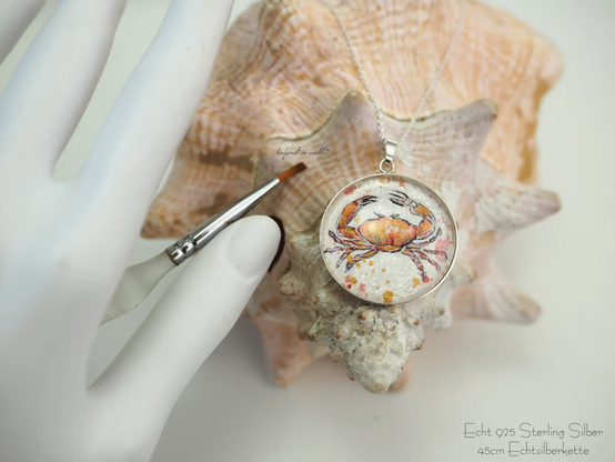 Handgemalte Krebs-Illustration, 30mm Durchmesser, Originalaquarell auf schimmernd perlmuttweiß grundiertem und gesprenkeltem Echtbüttenpapier, gefasst unter Glas und in Fassung aus Echtsilber festmontiert, auf Muschel fotografiert
