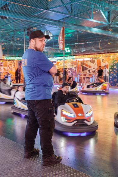 Ein Mann in einem blauen T-Shirt mit der Aufschrift "CREW" auf dem Rücken steht mit verschränkten Armen auf einer Metallplattform eines Autoscooter. Im Hintergrund sind mehrere Autoscooter mit Fahrgästen zu sehen, die sich auf der Fahrbahn bewegen. Die Umgebung ist bunt beleuchtet und mit verschiedenen Dekorationen versehen.