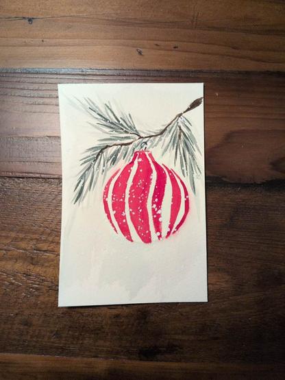 This is watercolor holiday card in portrait orientation on a 4x6 sheet of Arches Coldpress watercolor paper.  For this card I painted a branch of an evergreen tree with a peppermint inspired bauble hanging from it.  The painting is splattered with white paint to kind of give the appearance of snow.