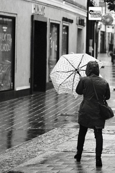 Das Foto zeigt eine Straßenszene in Schwarz-Weiß in der St. Pöltenter Innenstadt an einen regnerischen windigen Tag. Man sieht eine Person mit Regenschirm der durch den Wind verbogen ist.