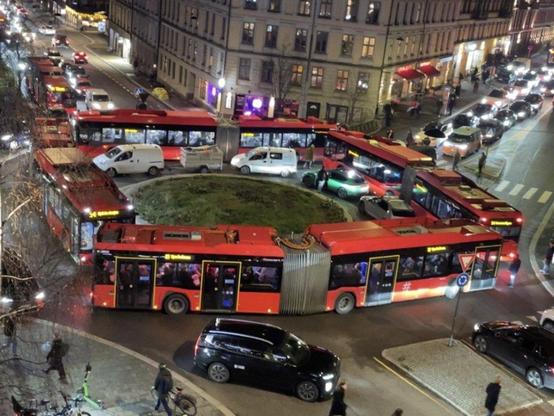 A scene showing four red bendy buses making up a squarw around a small roundabout. There are cars trapped inside them and long lines on the two visible sides going into the roundabout. Photo was found in Avisa Oslo, a local newspaper, and was sent them by 
a guy named Endre Helgeland.