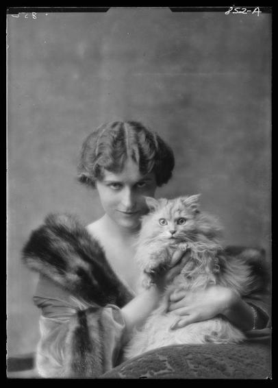 The image is a black and white portrait of a woman holding a fluffy cat. The woman has short, styled hair with bangs framing her forehead and cheeks. She appears to be wearing a dress or coat made from fur, as evidenced by the draped fabric on one shoulder and around the arms. Her expression is soft and gentle as she gazes directly at the camera.

The cat being held in her lap is large and fluffy, likely a breed with long hair such as a Persian or Maine Coon, based on its bushy coat and expressive eyes. The fur appears to be of various shades of cream or light yellow, adding texture to both subjects' presence in the photograph.

Both the woman's attire and the cat's grooming suggest that this image was taken during an era when fashion favored opulence and a sense of elegance, possibly early 20th-century based on her hairstyle and clothing style. The background is plain and nondescript, drawing focus to the subjects themselves. There are no discernible features or decorations in the backdrop.

The overall composition is intimate and seems personal, capturing a moment between the woman and her pet. It conveys warmth and affection towards animals as companions, which might have been considered significant during that time period. The photograph carries historical significance due to its vintage nature and portrayal of early 20th-century fashion trends in both human attire and animal grooming.