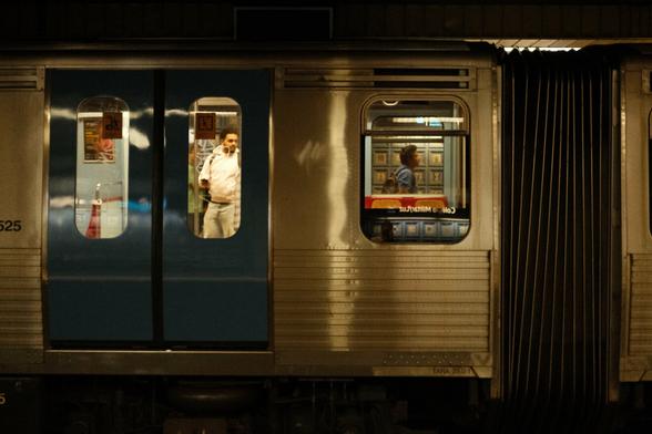 Picture of a subway train. More to the left, through the window of the closed door of the train you can see a person in white clothes standing or entering the train. To the right of that one, through the windows of the train, you see a person walking on the platform.