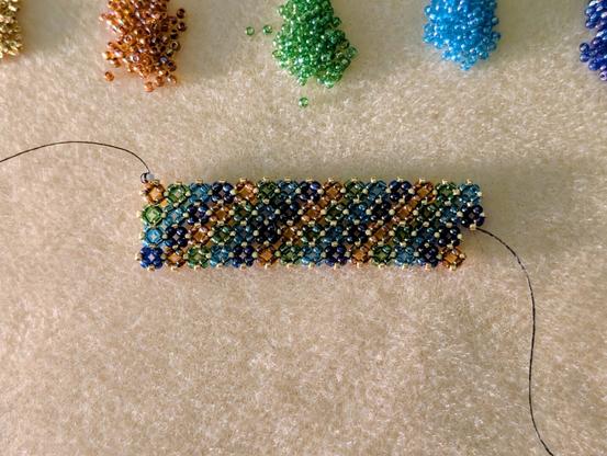 A partially finished beaded bracelet in orange, green, blue, purple, and gold seed beads. Above it are little piles of each color of beads that I'm working with.