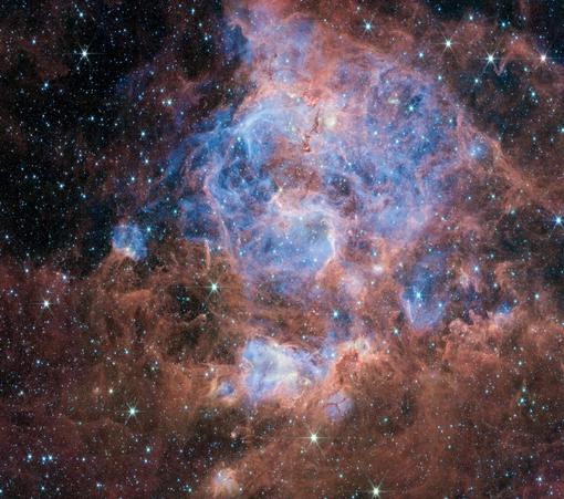 In this JWST image, a red and blue nebula spreads unevenly like a drifting cloud. The red forms an irregular, torn-fabric shape, while the blue pools in softer, rounded patches, like watercolor blooming on wet paper. Small stars and faint distant galaxies scatter across the dark background.