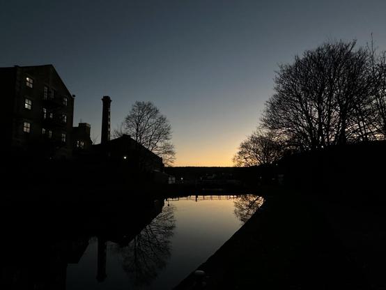 A view over a canal lock. There is an old mill building and a chimney on the left, and trees in the right. The last of the dusky orange sunset is reflected in the water, and fades into a grey blue sky above.