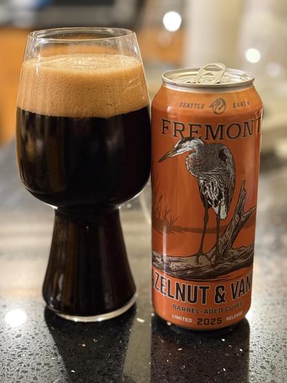 A stout glass is full of dark beer with a thick tan head. The orange can has black and silver accents and a long-legged tall crane or egret standing on a log in a pond