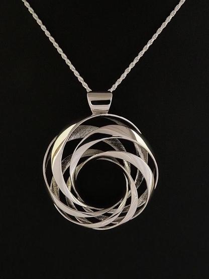 Photograph of Luminoso, a polished rhodium-plated brass pendant, about 35mm across, in the shape of an elegantly asymmetrical donut striped by six diagonal bands. The piece has a shiny inverted trapezoidal bail fitted with a silver rope chain and sits on a black background.