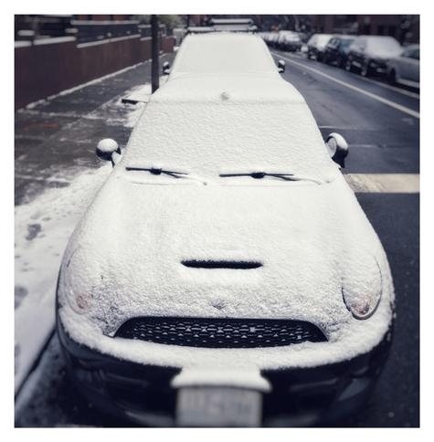 Photo of the front of a car parked on the side of a city street. It’s almost completely covered in a thin layer of snow, which contrasts with and highlights the windshield wipers, an indentation in the hood, and the front grill. These resemble facial features making a hilariously grumpy expression: the car seems to be glaring directly at the camera, with furrowed brows and gritted teeth.