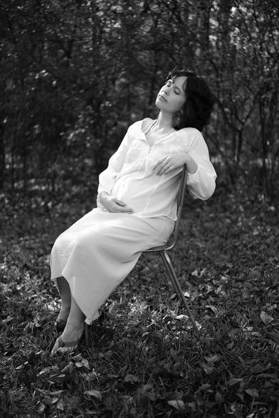 Black and white photo of a pregnant young woman. She's sitting in a chair, in her backyard, with her eyes closed and face lifted up towards the sunlight. She's cradling her baby bump with one hand. Fallen leaves cover the grass around her chair.