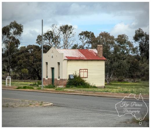 A small, historic looking building in Dumbleyung, Western Australia, with a distinctive design.  The front section is pale grey white with a small brick base and a peaked roof, while the main body is yellow cream corrugated metal with a rusty red corrugated iron roof and a brick chimney.  It sits near a paved road with a light post, surrounded by dry grass and scattered trees under a dramatic, cloudy sky.