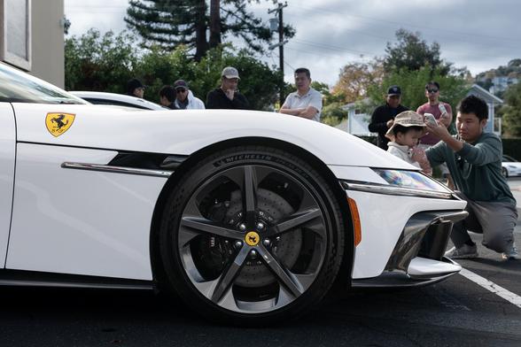 Front end detail of a white exotic’s long hood. A Ferrari crest is on the fender. People mingle in the background.