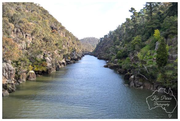 A photograph signed by Kev Peirce showing the Tamar River flowing between two steep, rocky slopes covered in dense green and scrubby brown vegetation.

The river water is a deep blue green, slightly rippled, and fills the narrow gorge. The banks are lined with dark boulders and rocks. The sky visible above the hills is bright white.