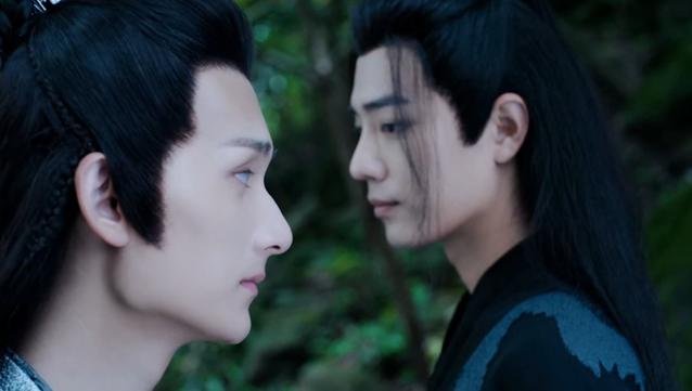 Close in shot of adult Wei Wuxian and Nie Huaisang against a green background of out of focus trees. Nie Huaisang and Wei Wuxian are standing side by side while facing opposite directions, with somber expressions following a warning delivered (Wuxian) and received (Huaisang.)