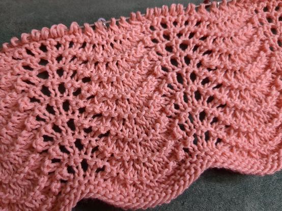 A section of an incomplete peachy-coloured knitted baby blanket. There is a small garter stitch border at the bottom, and then several repeats of the four-row Old Shale lace pattern, creating a rippled effect.
