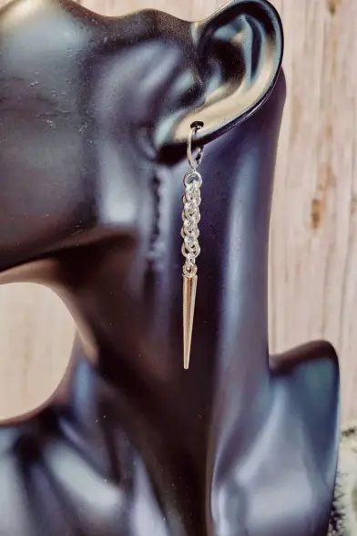 Boucles d'oreilles en maille composées d'une petite longueur de chaîne qui donne un effet de torsade et, au bout, d'une pique en acrylique. L'ensemble est couleur métal neutre.