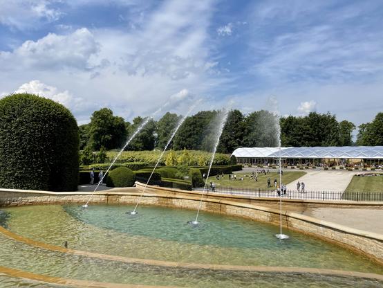 A landscaped garden features a tiered circular fountain with clear water and four arcing jets spraying into the pool. Behind the fountain, neatly trimmed hedges and topiary lead to a wide lawn where visitors walk and relax in small groups. A long, white, tent-like pavilion stretches across the background, with people gathered near its entrance and along the paths. The scene is framed by tall trees and a partly cloudy sky, creating a structured but open outdoor setting.