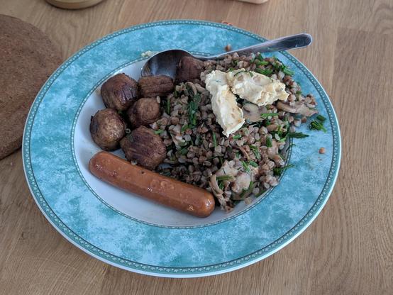 Auf dem Teller befindet sich ein herzhaftes veganes Gericht mit mehreren Komponenten:

Die Basis bildet gekochter Buchweizen, der mit Frühlingszwiebeln und Champignons vermengt wurde – eine aromatische Kombination, die sowohl nussige als auch frische Noten vereint.

Obenauf ist ein Klecks Hummus zu sehen, der dem Gericht eine cremige, würzige Komponente verleiht.

Als Beilage gibt es mehrere vegane Frikadellen, außen gut gebräunt und knusprig.

Ergänzt wird das Ganze durch ein veganes Würstchen, leicht gebräunt und servierfertig.


Das Gericht wirkt nahrhaft, proteinreich und ausgewogen – perfekt für eine sättigende vegane Mahlzeit.