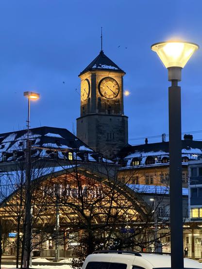 Ein beleuchteter Uhrturm in der Dämmerung mit zwei großen Zifferblättern. Im Vordergrund wirft eine Straßenlaterne ein warmes Leuchten, und ein Baum mit nackten Zweigen ist sichtbar. Schnee ist auf den Dächern der Gebäude in der Nähe sichtbar, was zur Winteratmosphäre beiträgt