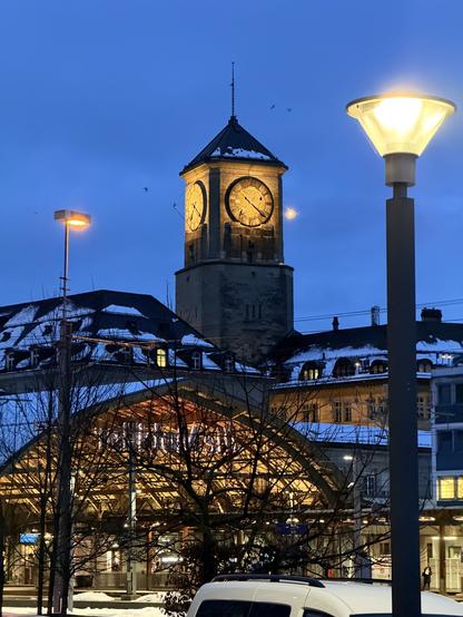 Ein beleuchteter Uhrturm in der Dämmerung mit zwei großen Zifferblättern. Im Vordergrund wirft eine Straßenlaterne ein warmes Leuchten, und ein Baum mit nackten Zweigen ist sichtbar. Schnee ist auf den Dächern der Gebäude in der Nähe sichtbar, was zur Winteratmosphäre beiträgt