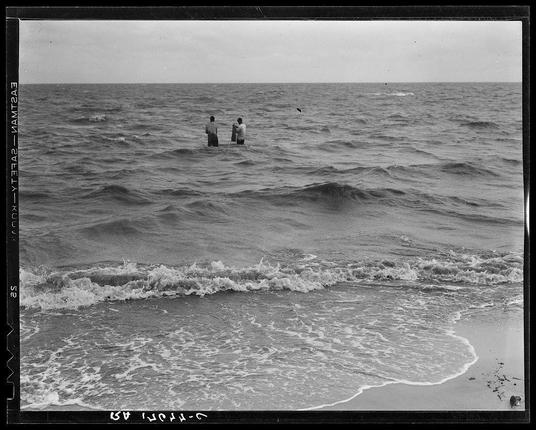 The image depicts three individuals standing waist-deep in the ocean. They are likely engaging in a leisure activity, possibly fishing or enjoying the sea. The scene is captured from an elevated perspective near the shoreline where gentle waves crash onto the sandy beach. This black and white photograph suggests it may be historical, as indicated by the presence of handwritten annotations along the left margin that appear to provide descriptive details such as "Gulf Coast" and a date range spanning over several months in 1935-6. The lack of modern equipment or attire could imply an earlier time period when sea-based activities were perhaps more commonplace for daily recreation rather than commercial endeavor, although fishing is suggested by the context provided.