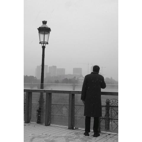 Black and white street photography taken in Luxembourg. A man in a dark coat, standing on the the corniche next to a lamp post stares at the skyline of the Kirchberg. Light snow is in the air and on the ground.