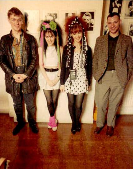 Four people stand in a room against a wall, smiling. Rose Mcdowall and Jill Bryson are in the middle in short dresses and hairbands with plastic flowers and long trailing ribbons. They are flanked by two casually dressed men of a comparable age. The walls have pictures of musicians, it appears to be one of Ringo Starr just behind Jill.