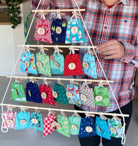 Eine Person im karierten Hemd hält einen selbstgemachten Adventskalender hoch. Der Adventskalender besteht aus Holzstäben und weißer Kordel und hat die Form eines nach oben spitz zulaufenden Tannenbaums. An den Holzstäben sind Stoffsäckchen in den Farben Grün, Rot und Blau mit Holzklammern befestigt. An jedem Stoffsäckchen hängt ein runder Holzanhänger mit einer Zahl.