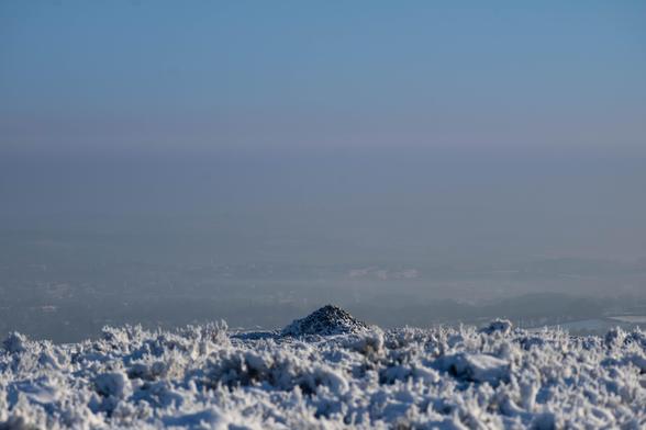 A cairn of grey stones on a snow covered moor, overlooking lower land on the edge of the city. Above, blue skies
