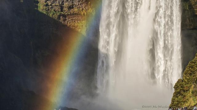 A colour photo of most of a waterfall dropping directly in a curtain of white water in front of a rockface. The spray has created a strong rainbow on the left. There are people standing at the sides of the falls, only partially seen.
