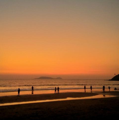 Silhouettes of people stand on a beach at sunset, with a vibrant orange sky and a calm ocean. An island is visible on the horizon.