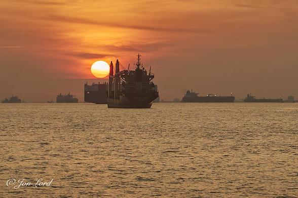 This is a seascape photo of a rising sun over the Singapore Strait with numorous ships at anchor. Singapore (2017).

The surface of the sea is calm with only the smallest wavelets breaking the surface with has an orange or golden colour reflected from the just risen sun. In the centre of the image is the stern of an anchored, medium sized, unlaiden container ship. The hull is almost in line with the camera with the port side of the hull in view with two cranes attached to that side of the ship. Forward of this ship is a car carrier, a blunt, flat sided and ugly ship that is anchored at right angles to the camera and partially obsured by the first ship. To the left of the container ship and above the car carrier is the large golden orb of the rising sun, just a few degrees above the horizon. Along the horizon are ten to fifteen large cargo ships either at anchor or underway through the Singapore Strait. Above the horizon is an orange or golden sky, made more colourful by a thin veil of cirrus clouds.