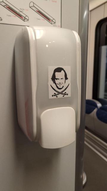 Adesivo attaccato in un dispenser per igienizzante su una carrozza Trenitalia.  Volto 2D stencil di Jack Nicholson leggermente modificato (logo di #no_adesivigrazie) sopra due spade incrociate (atte ricordare la bandiera dei pirati caraibici).
