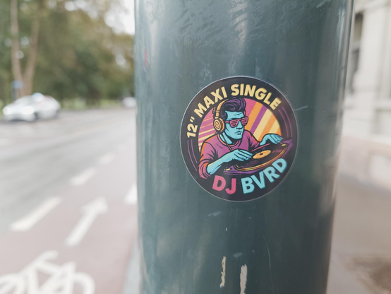 A colorful circular sticker on a dark pole features a DJ in sunglasses and headphones spinning a record. Text reads "12" MAXI SINGLE DJ BVRD".