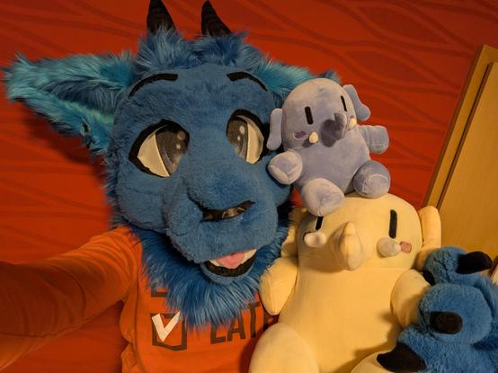 A selfie of me in my blue dragon partial fursuit, having the big yellow plushtodon in my paw. A smaller, blue colored plushtodon sits on the head of the big one.