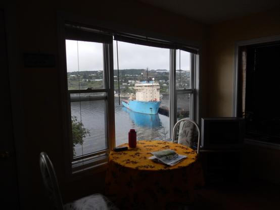 Picture looking across a dark room out a large window. There is a table with yellow table cloth, and two chairs in front of the window, and the view is of a ship with a blue hull in a harbour.