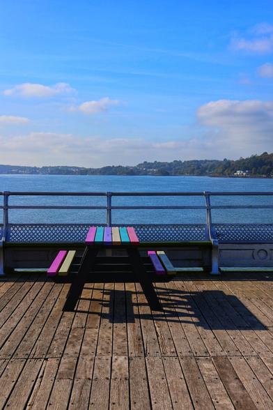 A vibrant, colourful picnic bench sits on a wooden pier overlooking a serene body of water. The bench features rainbow-coloured slats in shades of pink, yellow, green, blue, and purple, contrasting with its black metal frame. The pier, constructed from wooden planks, extends towards a metal railing with decorative lattice panels. Beyond the railing, calm waters stretch towards a distant shoreline lined with trees and scattered buildings. The sky above is clear and blue, dotted with a few fluffy white clouds, creating a peaceful and inviting atmosphere.
