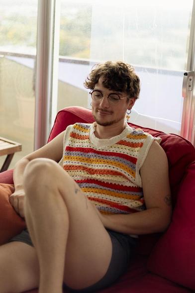 Quelqu’un est affalé dans un fauteuil rouge, tranquille, comme si la journée le laissait enfin respirer. Son pull sans manches, bariolé de rayures chaudes, ajoute une petite note artisanale.