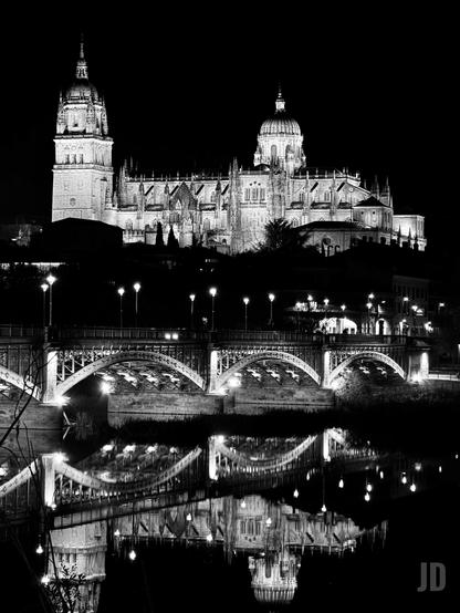 Imagen nocturna en blanco y negro de la Catedral de Salamanca y el Puente Enrique Estevan sobre el río Tormes. La majestuosa catedral, con su alta torre con chapitel a la izquierda y una gran cúpula central, está intensamente iluminada y se alza contra un cielo completamente oscuro. Debajo, el puente de piedra y metal con múltiples arcos, también iluminado por farolas, cruza el río. Las luces de la catedral y el puente se reflejan nítidamente en la superficie del agua, creando una imagen simétrica y serena.