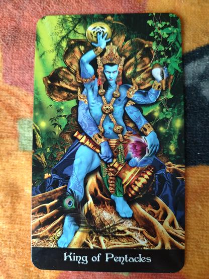 Card from the Tarot Apokalypsis deck. 
King of Pentacles.
A male god of blue skin, tattooed, a man with four arms. In each hand he holds an object: a pentacle, a conch, a large scepter, and a flower. He sits on a throne in the woods.