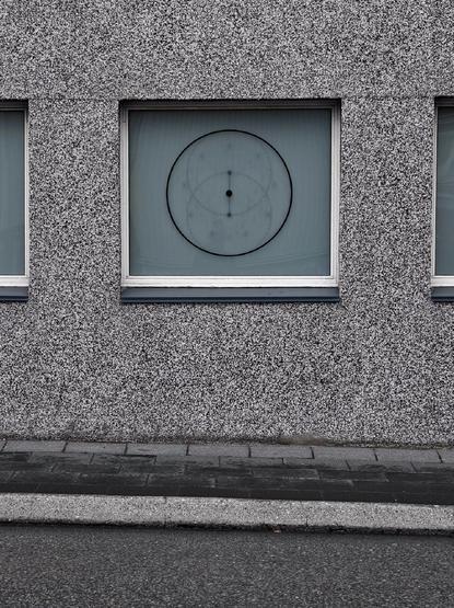 Colour photo showing a grey outside wall above a grey street. In the middle is a frosted window displaying a large circular pattern with a fainter pattern inside and small black dots at the center.