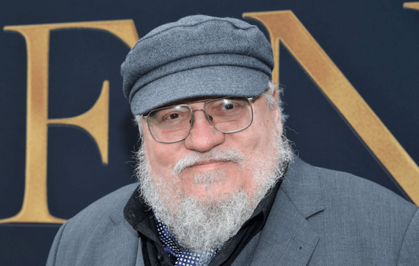 Un homme avec une barbe grise et des lunettes sourit à la caméra tout en portant une casquette grise et un costume bleu, devant un fond orné du mot 'FEN'.