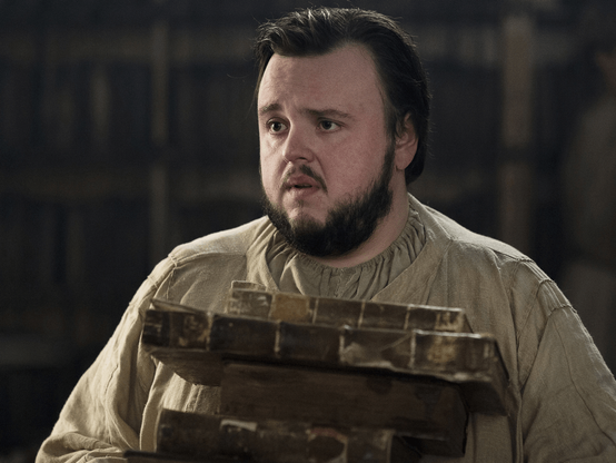 Un personnage portant des vêtements simples et un air préoccupé, tenant plusieurs livres en bois dans une bibliothèque sombre, avec des étagères remplies de livres en arrière-plan.